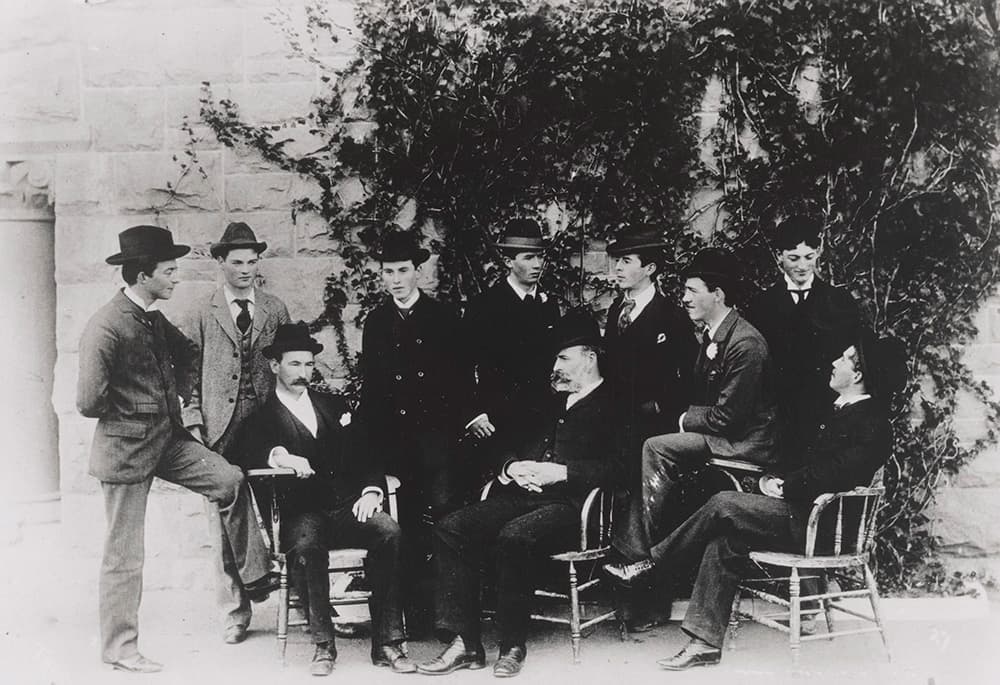 Black and white photo of nine men in vintage suits and hats; some seated and others standing against a stone wall with ivy.