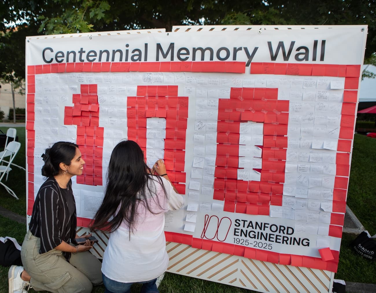 Two people add notes to a Centennial Memory Wall filled with red and white sticky notes forming the number 100 for Stanford Engineering's 1925-2025 celebration.