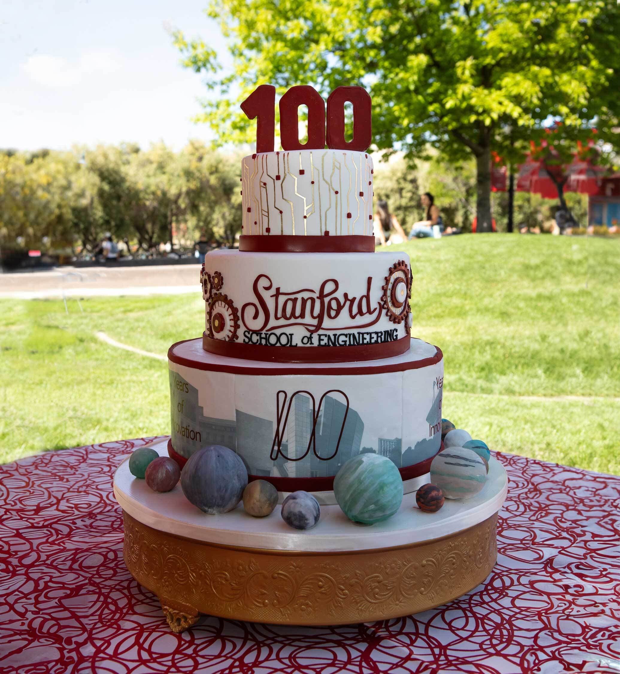 Four-tier white cake with 'Stanford School of Engineering' written in frosting on the side and candles shaped like the number 100 on top.