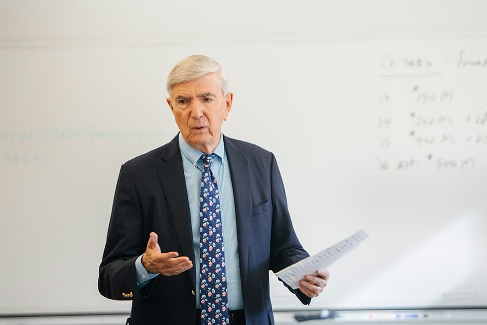 A man in a suit and tie stands in front of a whiteboard, holding papers and gesturing as he speaks.
