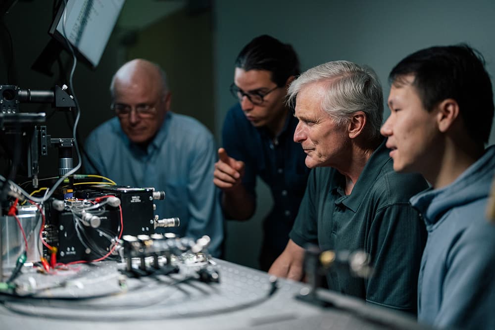 Four men intensely focus on a complex machine with wires and screens, exuding curiosity and teamwork in a dimly lit lab setting.