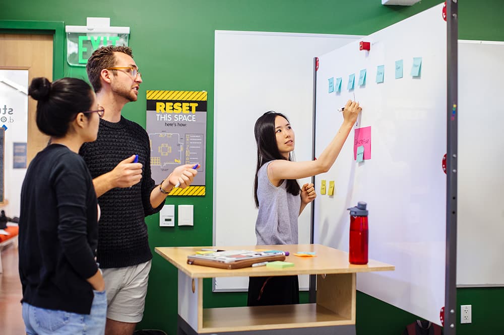 A group of three people collaborates in a bright room. One person writes on a whiteboard covered with sticky notes, while the others watch intently.