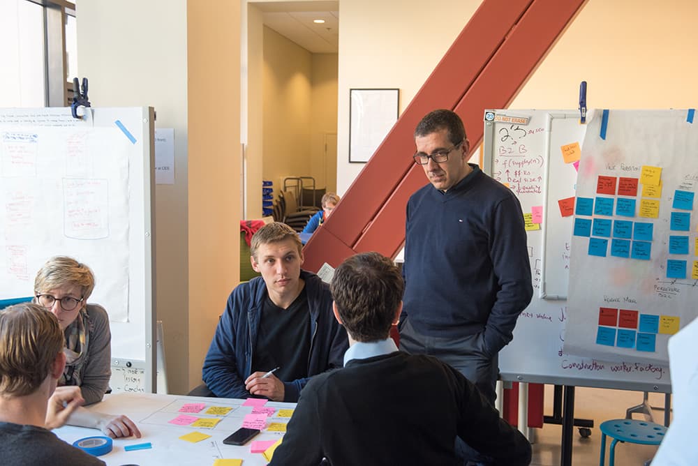 A group collaborates around a table covered with sticky notes and papers. A man stands, listening intently.