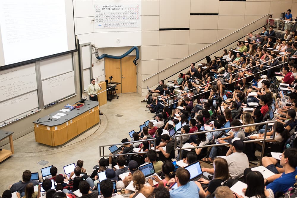 A professor teaches a large college lecture in a tiered classroom filled with students, many using laptops. A periodic table and whiteboards are visible at the front.
